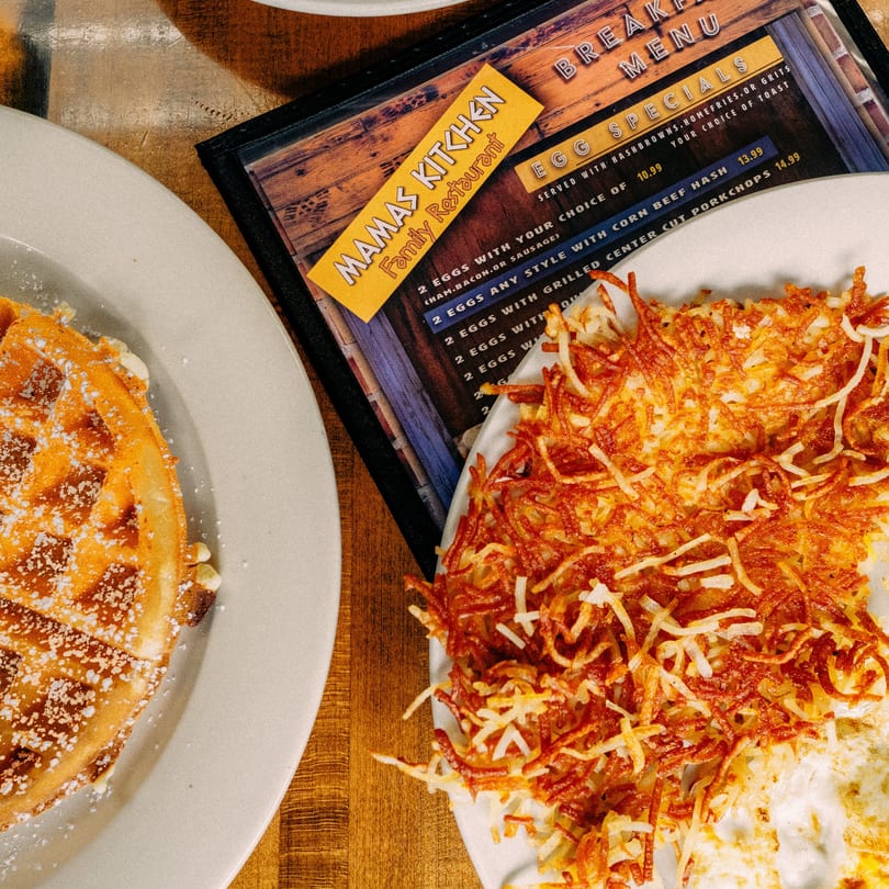 A waffle with powdered sugar and a plate of hash browns next to a Mamas Kitchen breakfast menu.
