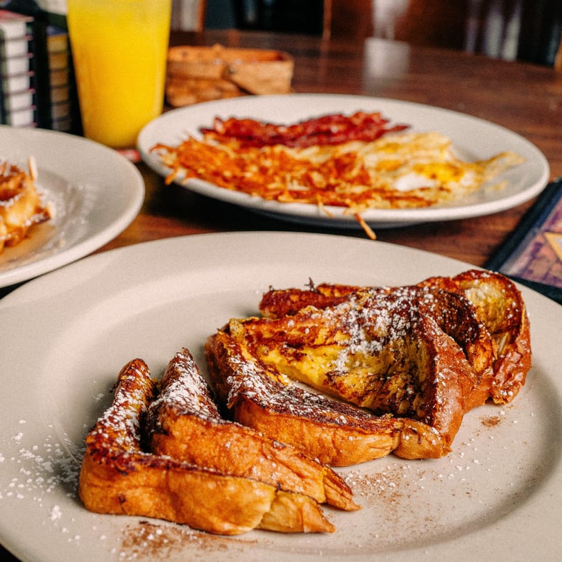Mamas Kitchen's French toast dusted with powdered sugar and cinnamon, with hash browns and bacon in the background.